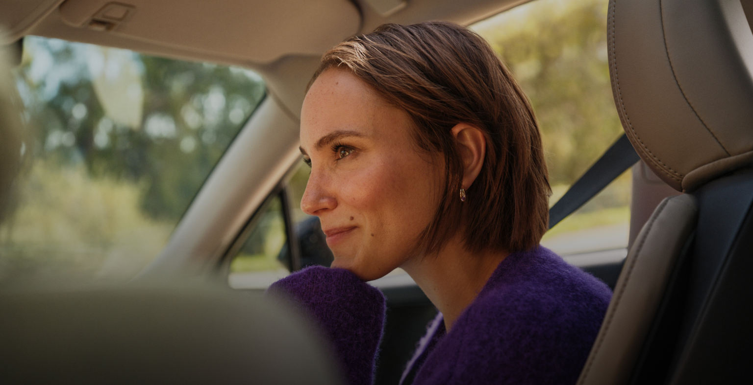 Smiling woman with earrings and purple straps holding her hair back against a clear blue sky.