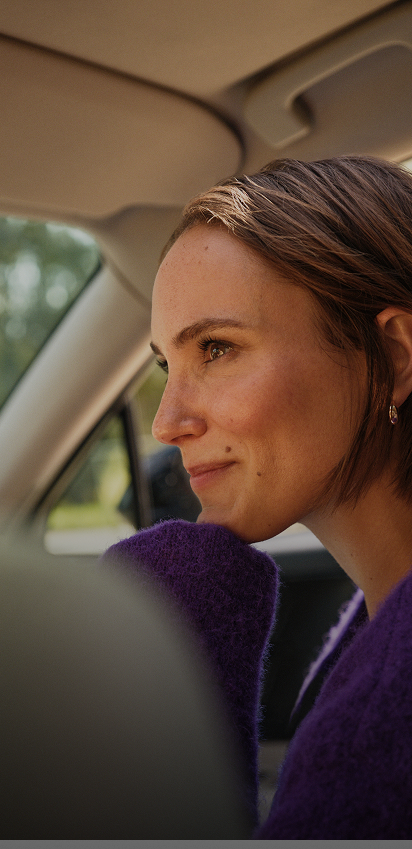 Smiling woman with earrings and purple straps holding her hair back against a clear blue sky.