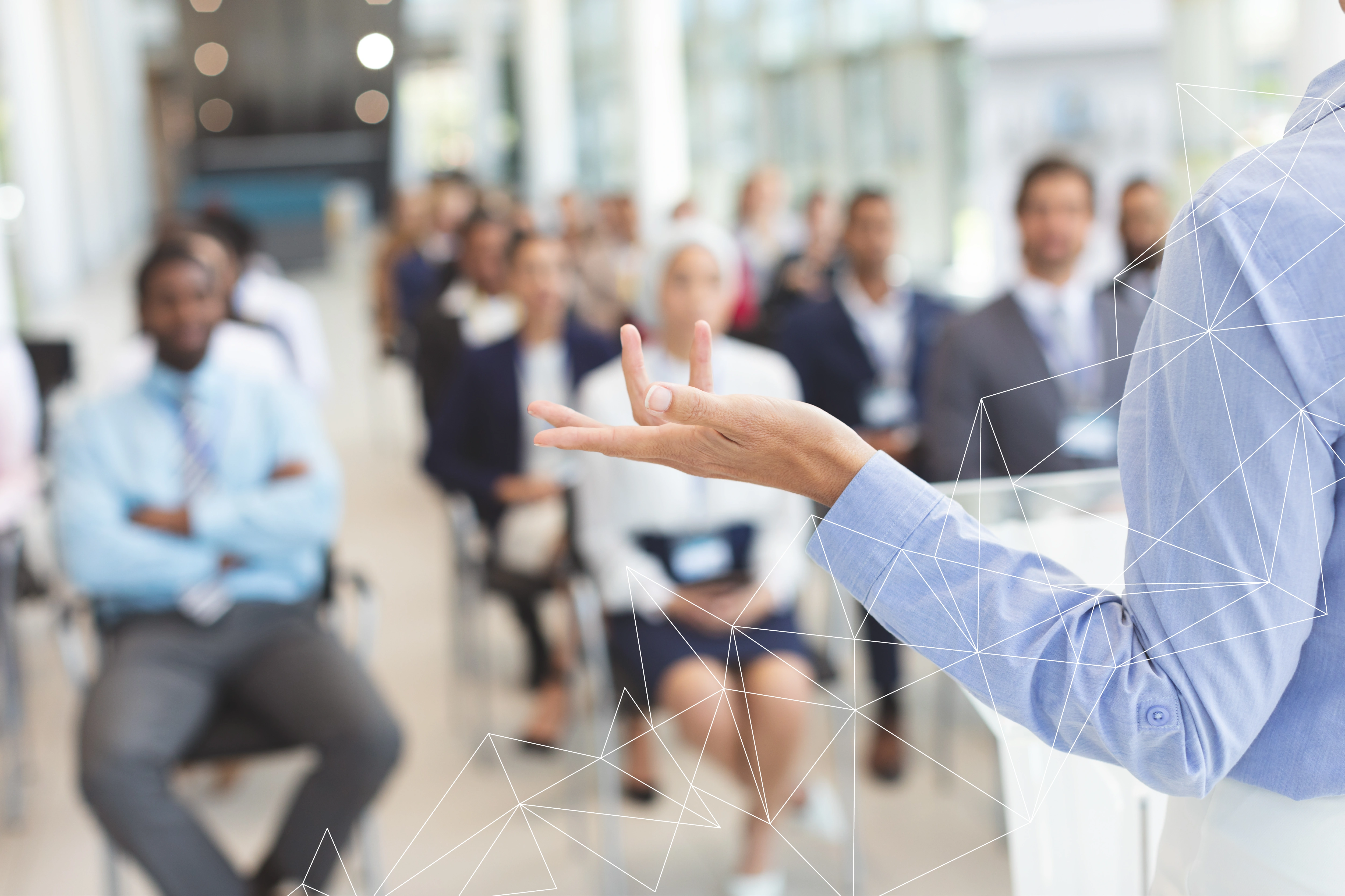 Speaker in blue shirt gesturing with hand to an audience blurred in the background.