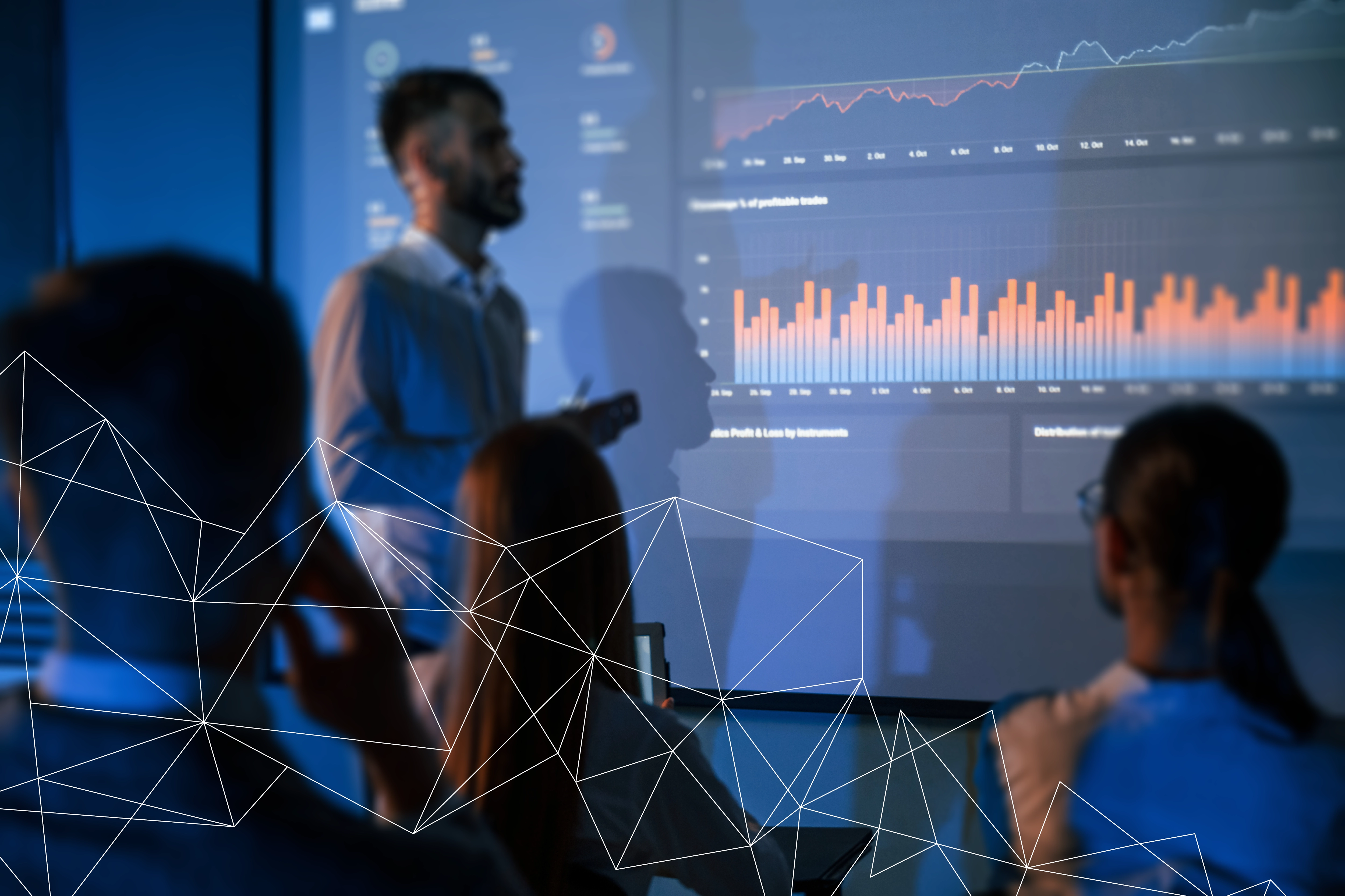 Man presenting financial data with bar and line charts to seated colleagues in a dimly lit room.