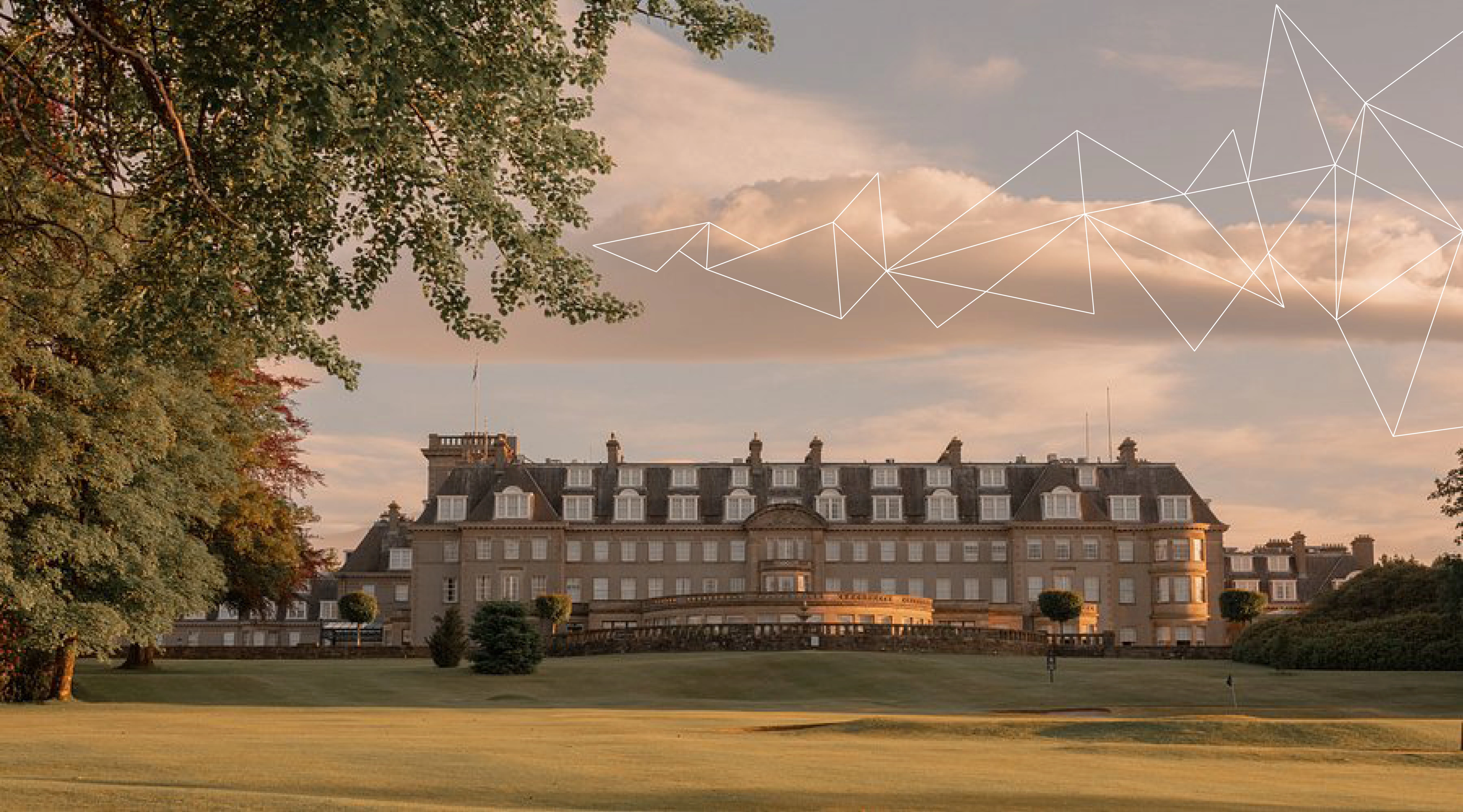 Large historic hotel building with many windows, surrounded by green lawn and trees at sunset.