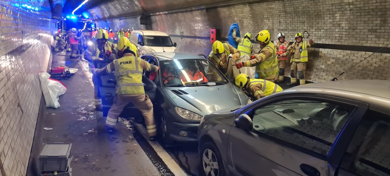 Firefighters in yellow helmets assist with a car accident rescue inside a tunnel with emergency lights active.