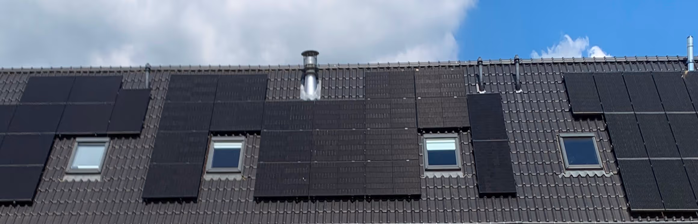 Roof with multiple black solar panels installed around three skylights under partly cloudy sky.