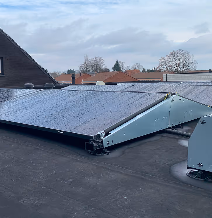 Solar panels installed on a flat rooftop with nearby houses and trees in the background under a cloudy sky.
