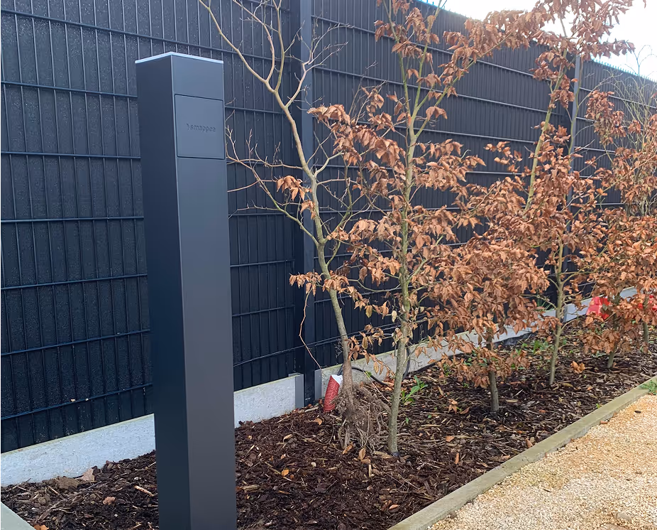 Tall black Smappee outdoor electric vehicle charging station next to a row of small trees with brown leaves and mulch ground cover.