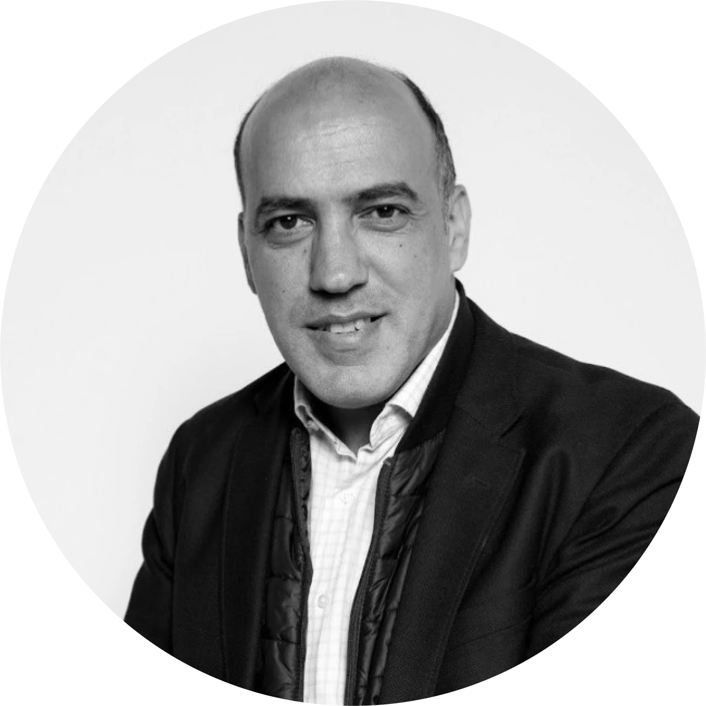 Black and white portrait of a smiling bald man wearing a dark blazer over a quilted jacket and collared shirt.