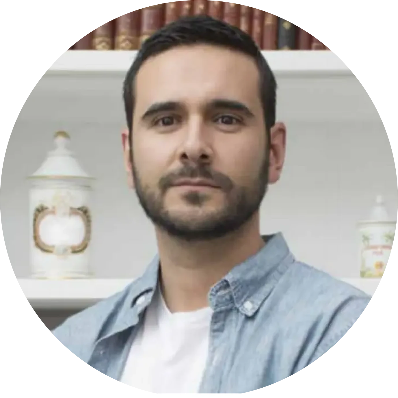 Portrait of a man with short dark hair and beard wearing a light blue denim shirt and white t-shirt in front of a bookshelf.