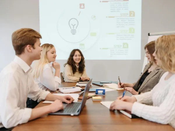 Group of five young professionals sitting around a conference table with laptops and notebooks, engaged in a meeting with a presentation projected on the wall.