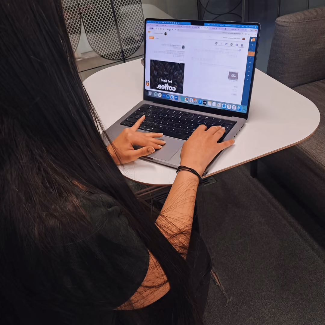 Person with long hair typing on a laptop at a white table in a casual workspace.