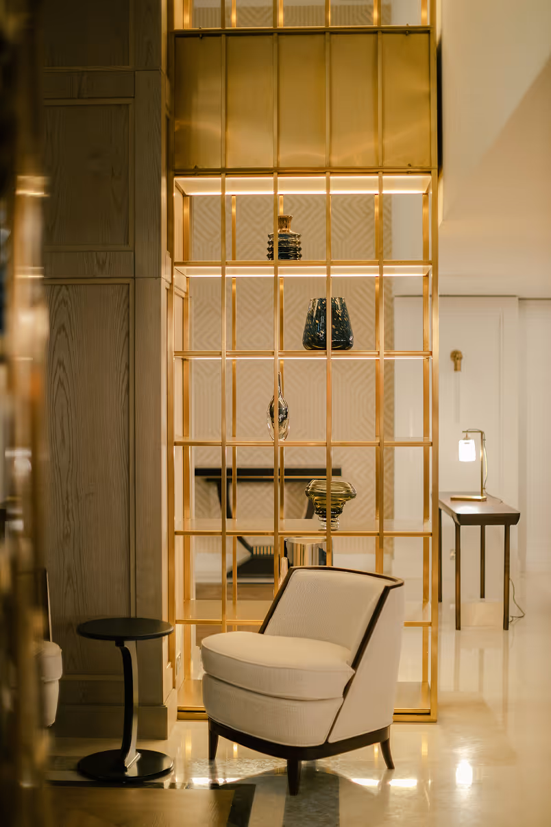 Modern sitting area with a white cushioned armchair, small black side table, and a gold metal frame shelf displaying decorative glass vases.
