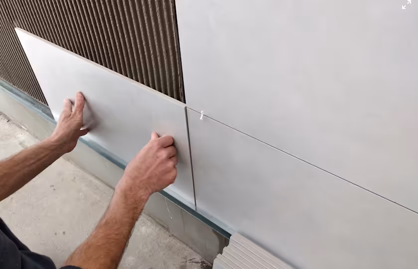 Person installing large gray wall tiles onto adhesive on an exterior wall.