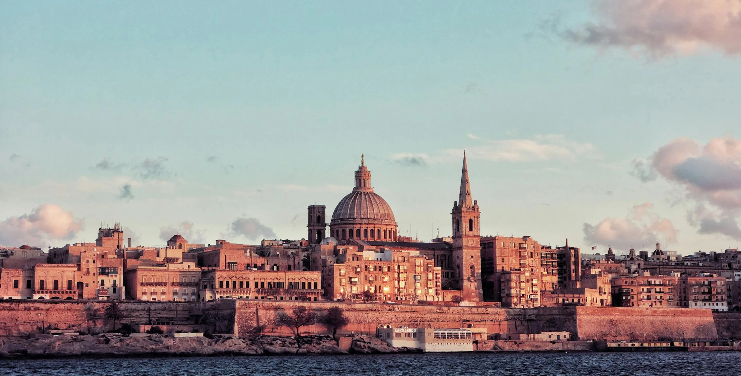 The image displays the iconic skyline of Valletta, the capital city of Malta, as seen from across the water. 