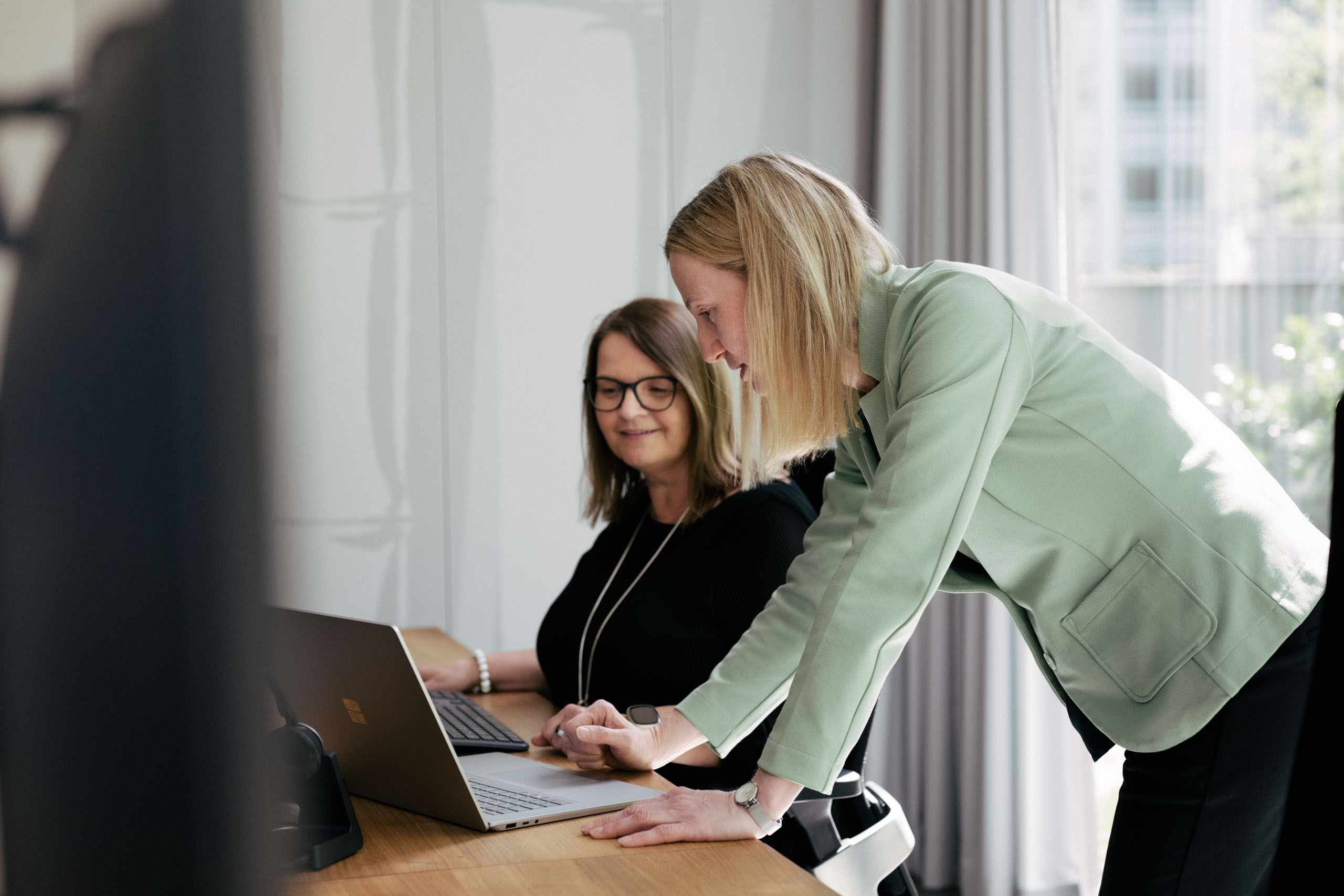 Zwei Frauen arbeiten gemeinsam an einem Laptop in einem hellen Büro mit großen Fenstern