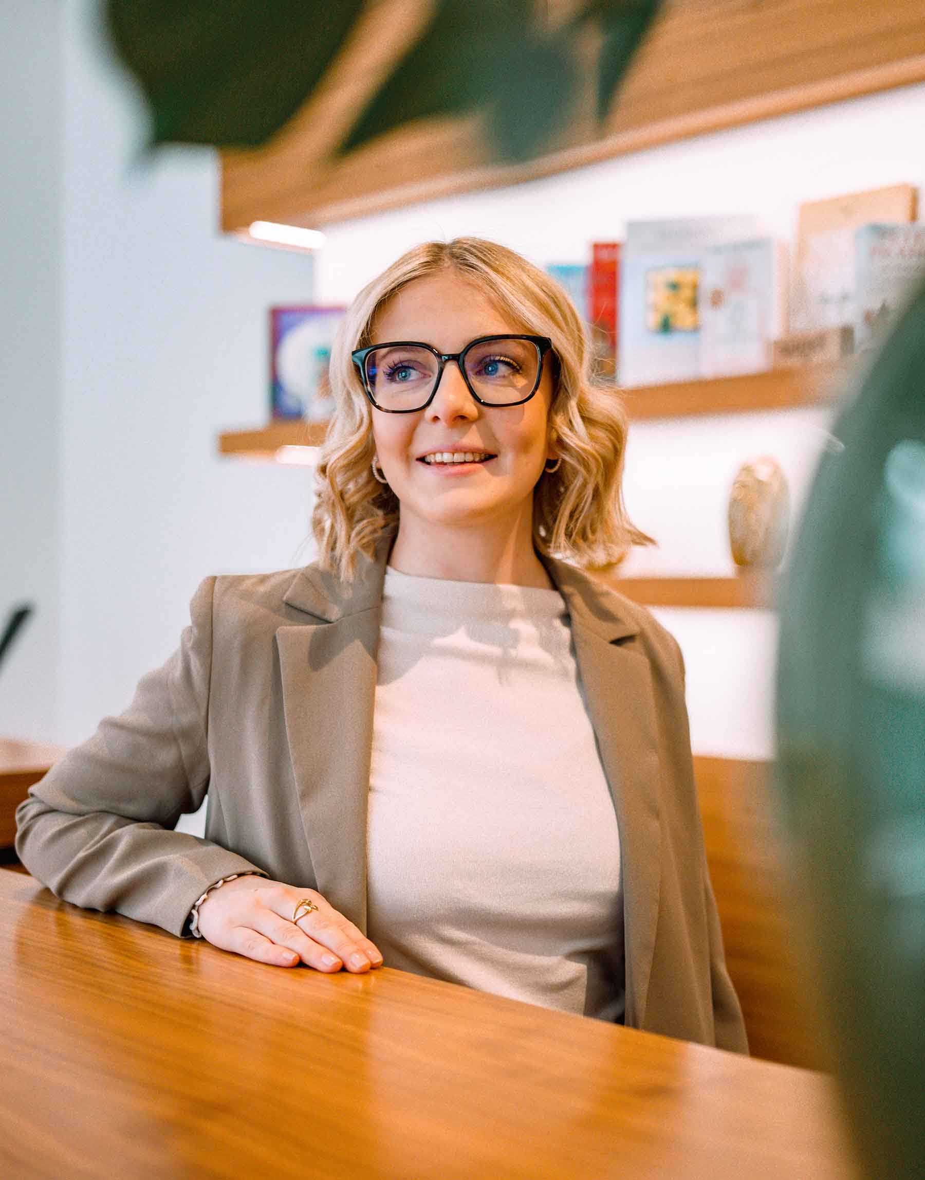 Zwei Personen unterhalten sich an einer hölzernen Theke in einem modernen Büro. Links steht eine blondhaarige Frau in schwarzem Blazer und weissem Oberteil, rechts ein junger Mann mit Brille und weissem Hemd, der eine Tasse Kaffee hält. Im Hintergrund sind Barhocker und eine Vase mit rosa Blumen zu sehen.