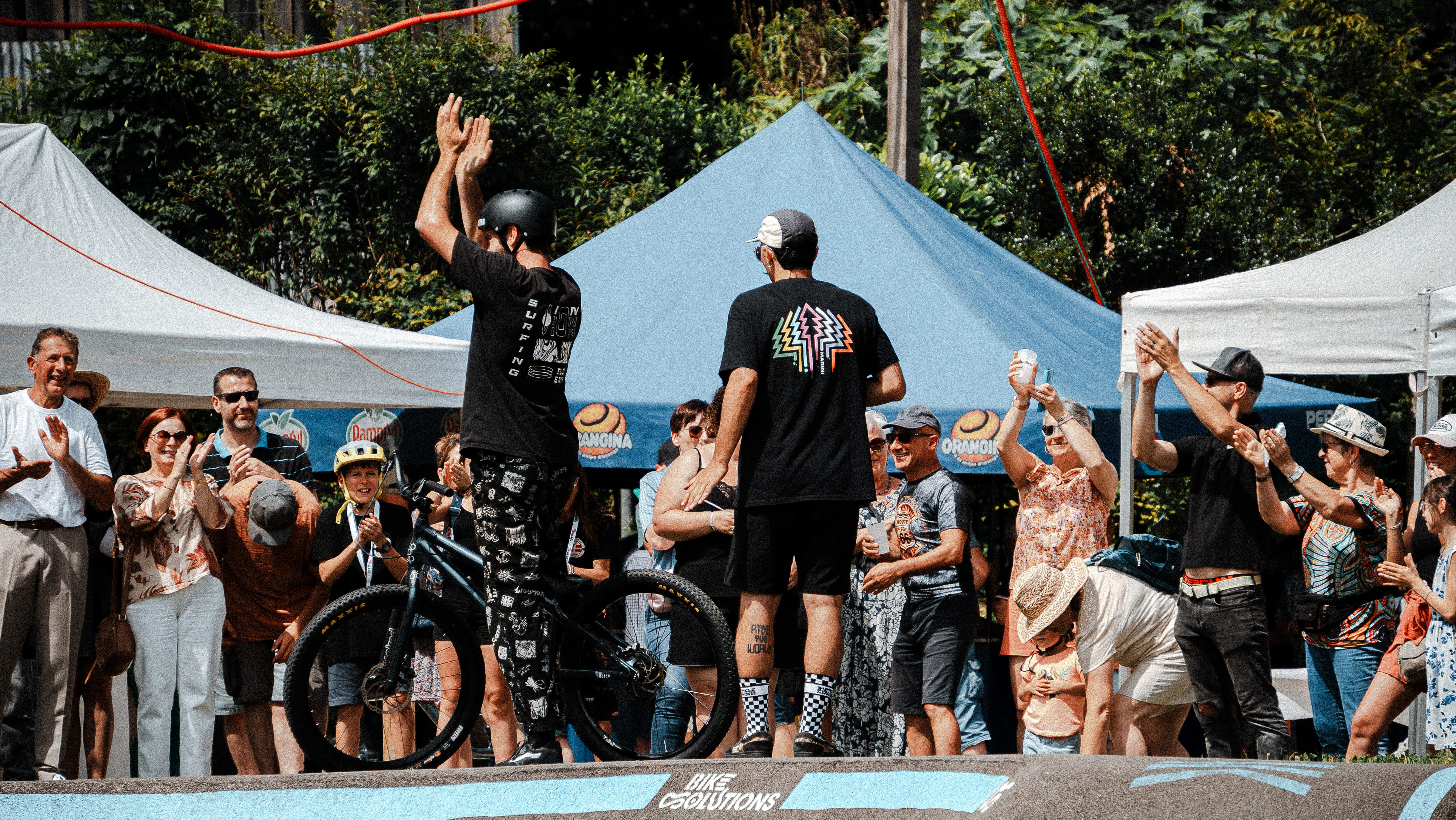 Bicyclist wearing a helmet and patterned pants standing on a pump track stage with hands raised while a crowd claps and cheers around him under blue and white tents.
