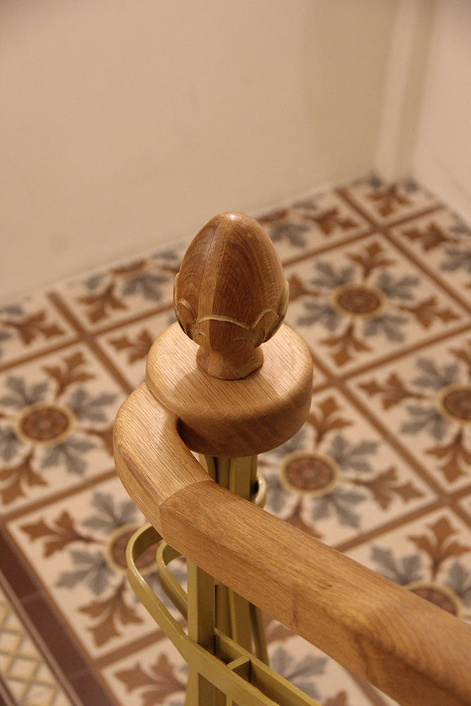Curved wooden handrail with decorative finial above patterned floor tiles in brown and gray tones.