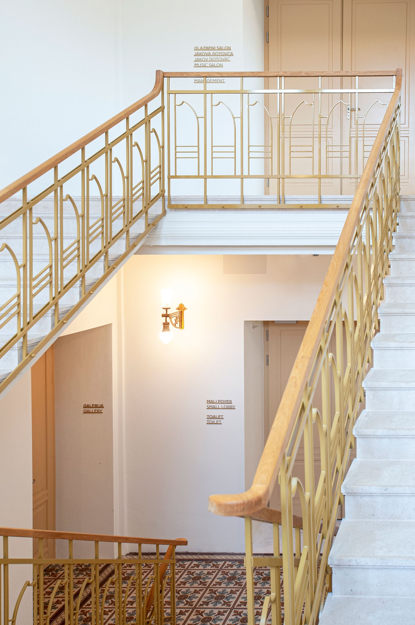 White staircase with beige railings and patterned floor tiles in a building interior featuring signs for gallery, music salon, small lobby, and toilet.
