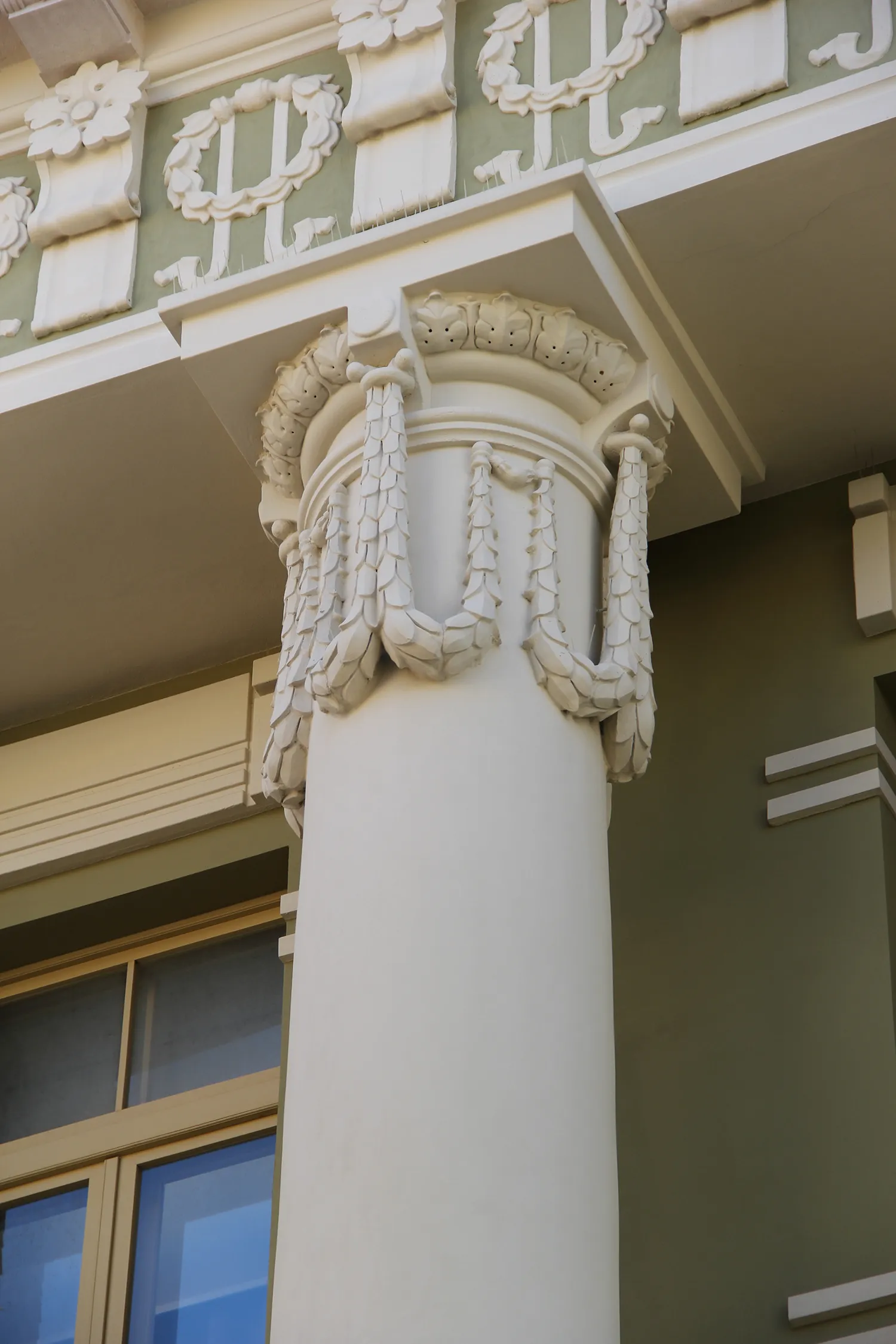 Ornate white column with sculpted leaf-like garlands and floral decorations near the top, set against a green building facade.
