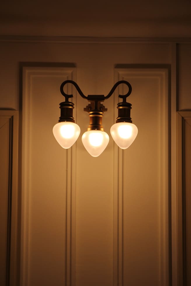 Three-bulb vintage wall sconce glowing warmly on an off-white paneled wall.