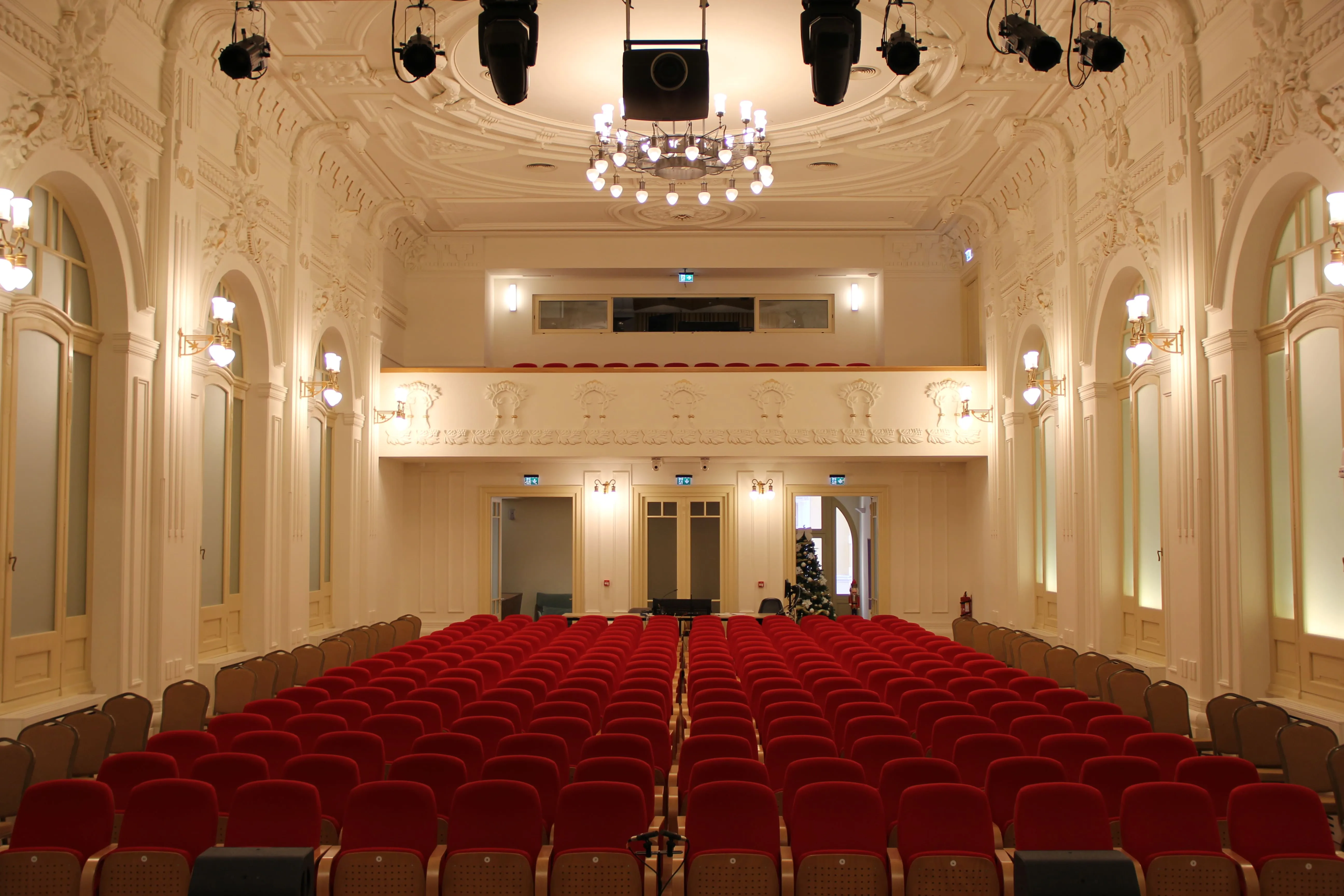 Elegant auditorium with red velvet seats, ornate white walls, and a chandelier hanging from a detailed ceiling.