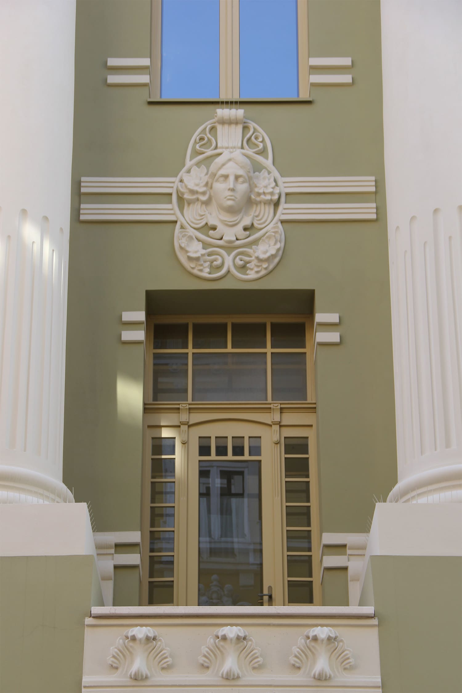 Ornate door with glass panels set in a green wall, flanked by white classical columns and topped with a decorative sculpted face.