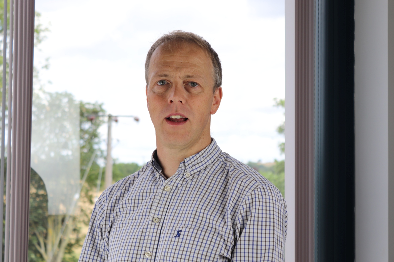 Man in a checkered button-up shirt standing indoors near a window with trees visible outside.