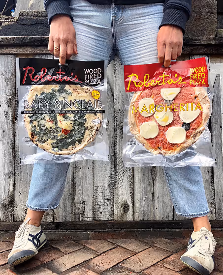 Person holding two packaged Roberta's wood fired pizzas, Baby Sinclair and Margherita, wearing light blue jeans and white sneakers.