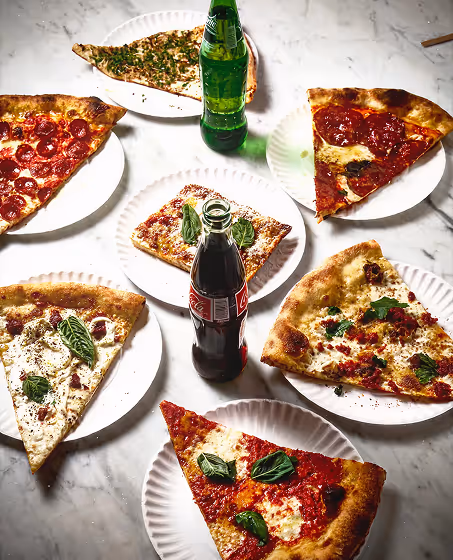 Six slices of assorted pizza with fresh basil leaves on white paper plates arranged around two glass bottles of soda on a marble table.
