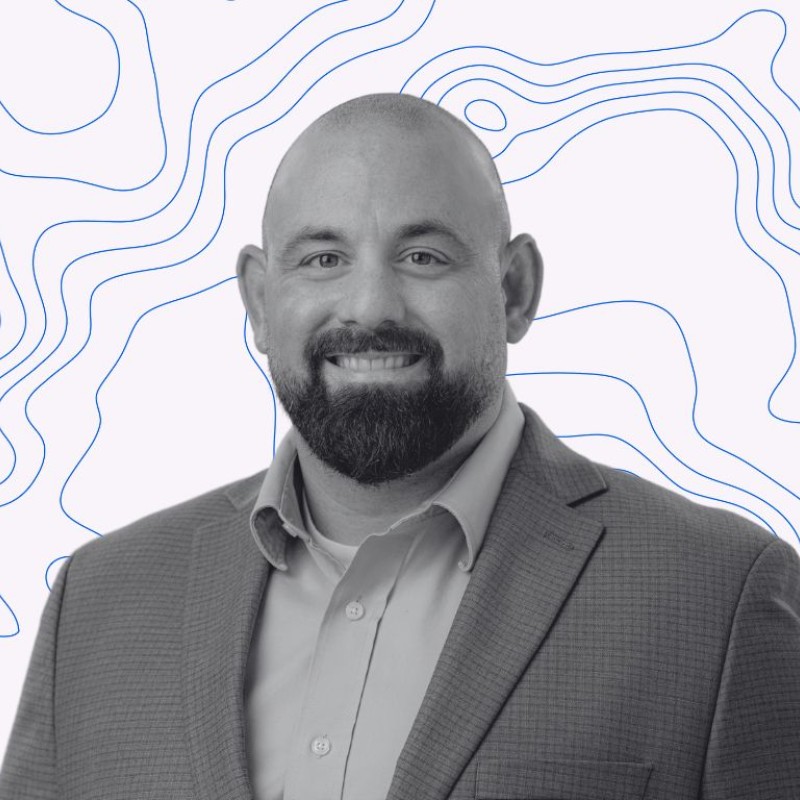 Smiling bald man with a beard wearing a suit jacket and dress shirt against a background with blue contour lines.