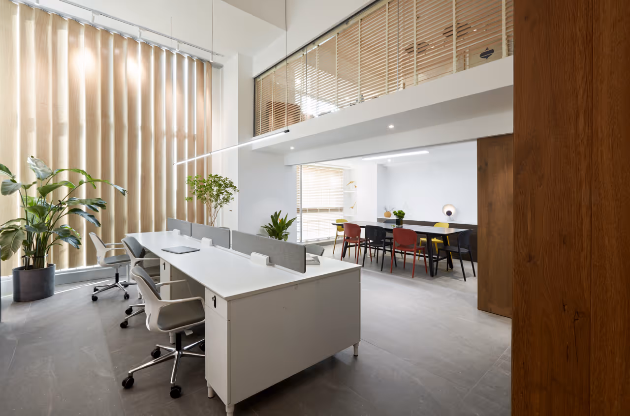 Modern kantoor met witte bureaus, draaistoelen, planten en een vergadertafel met kleurrijke stoelen.