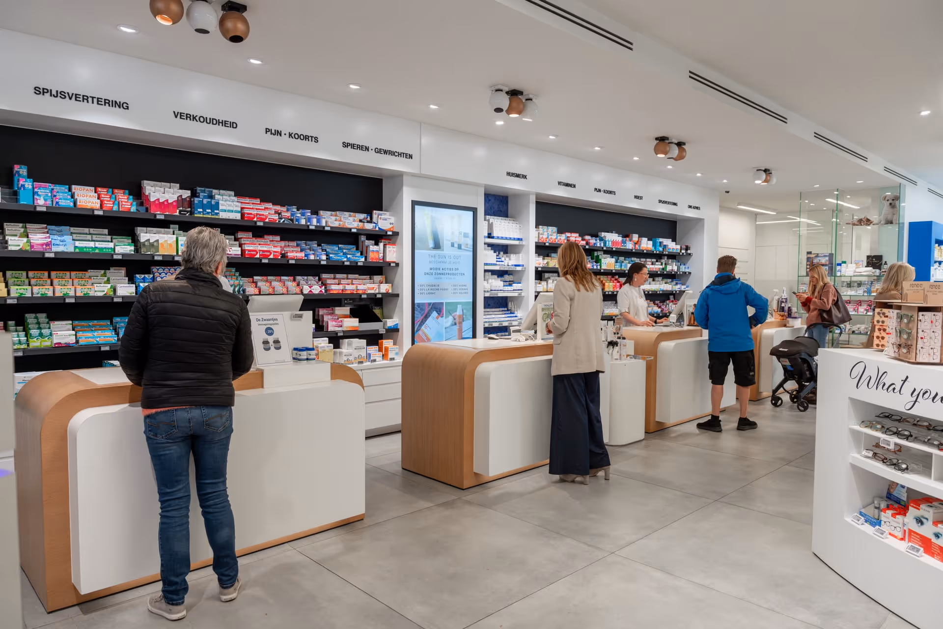Klanten aan de balies in een moderne apotheek met schappen vol medicijnen en gezondheidsproducten.