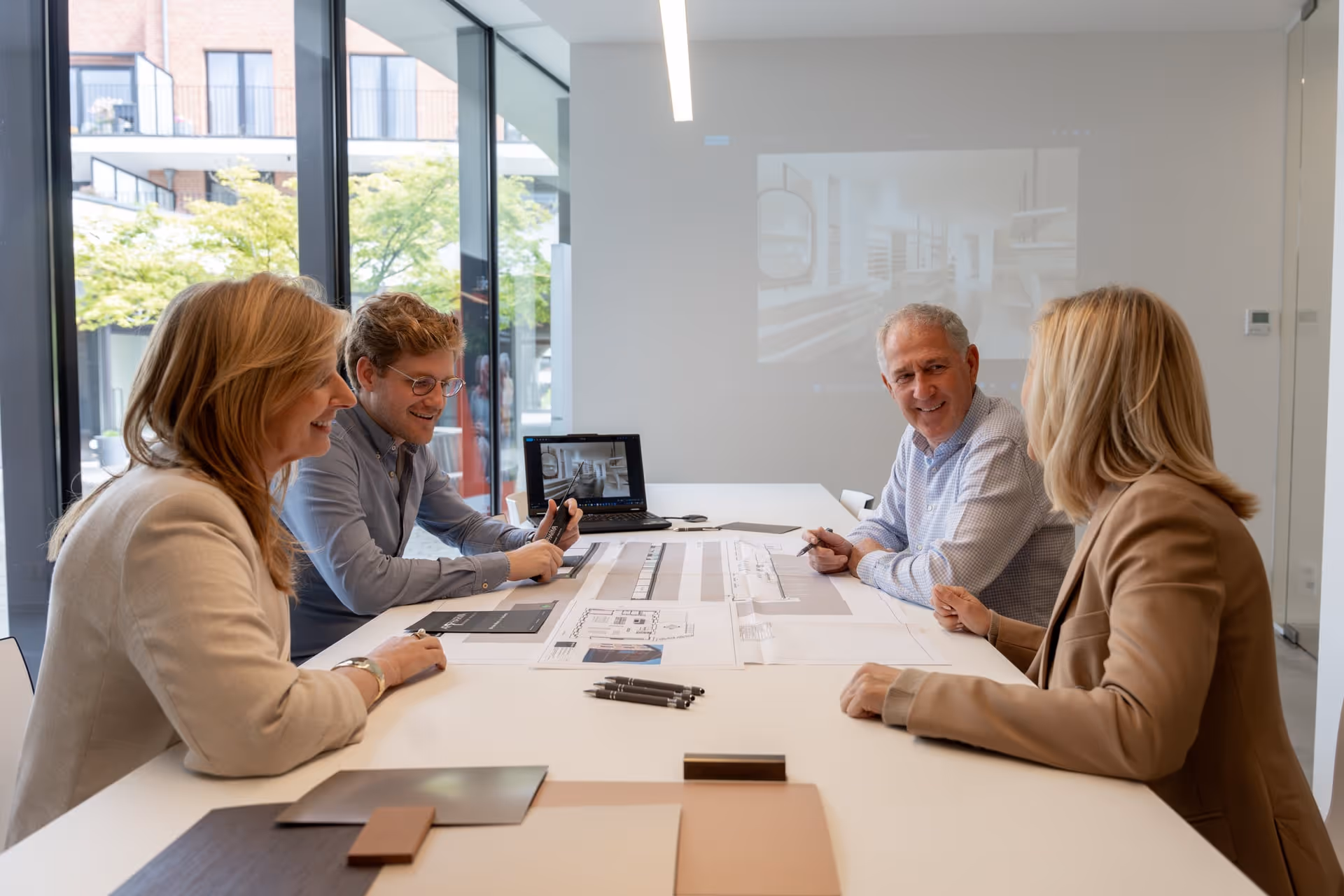 Vier mensen die rond een witte tafel in een modern kantoor met grote ramen over interieurplannen praten.