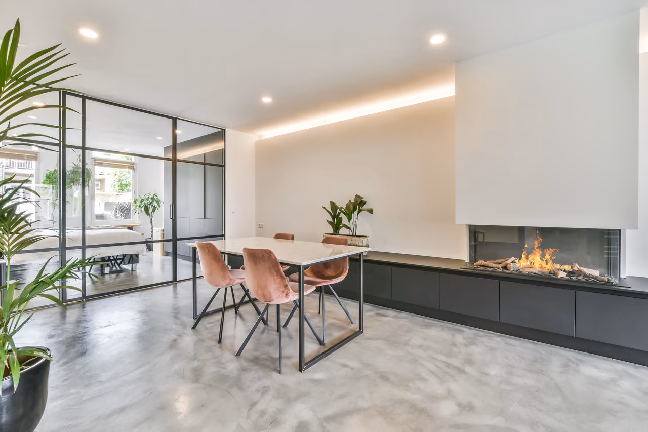 Moderne eethoek met roze stoelen, een glazen wand, potplanten en een brandende open haard op een gepolijste vloer.