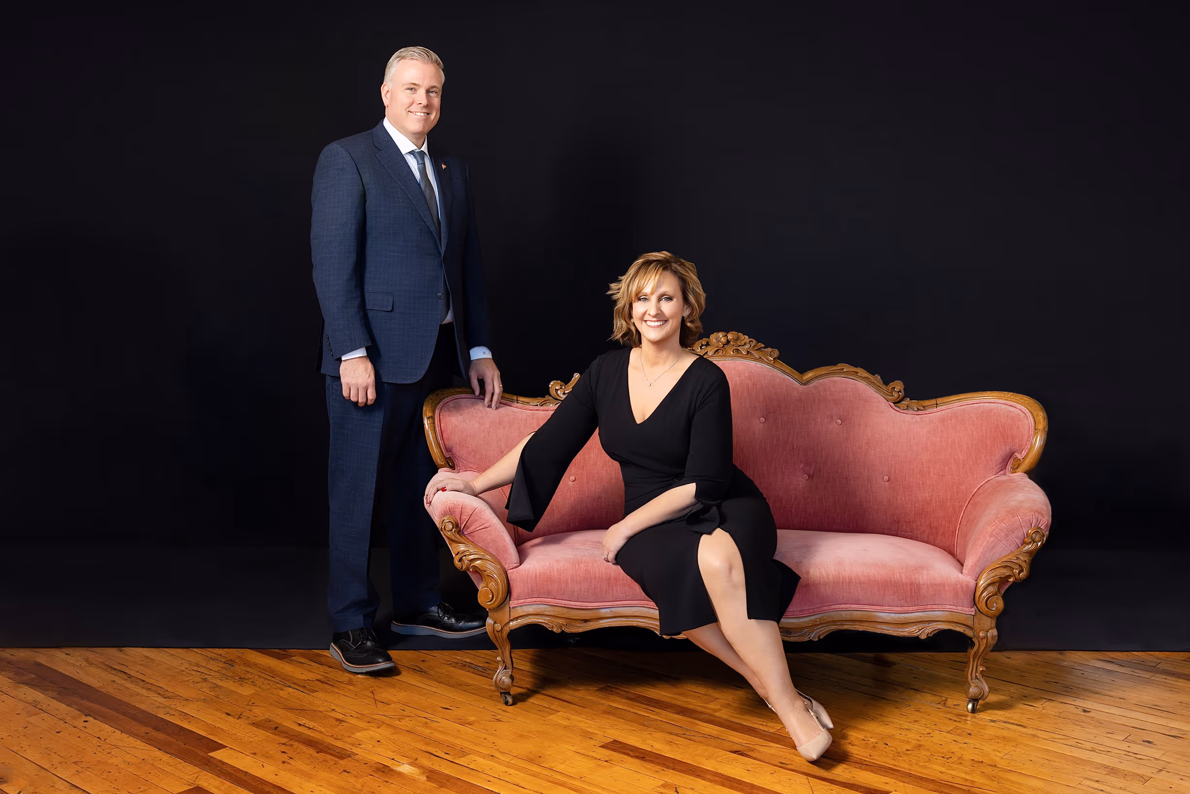 Man in a navy suit standing next to a woman in a black dress seated.