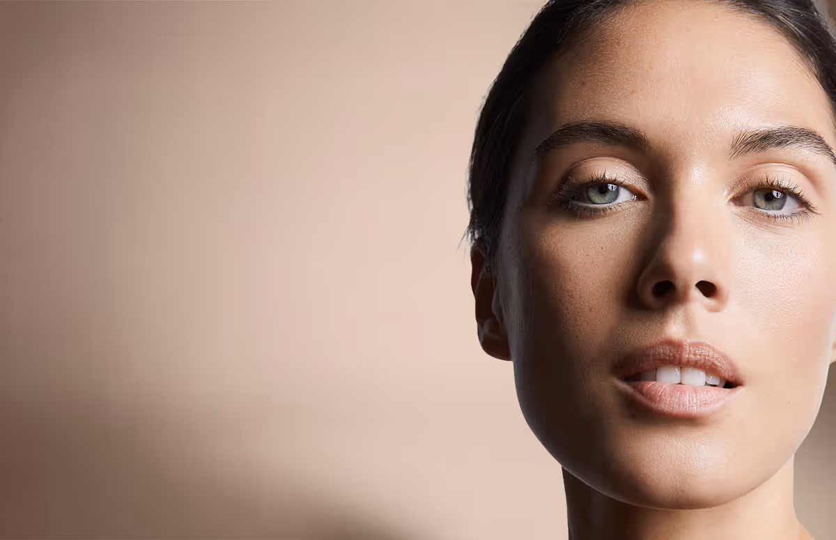 Close-up of a woman's face with clear skin and light makeup looking into the camera against a beige background.
