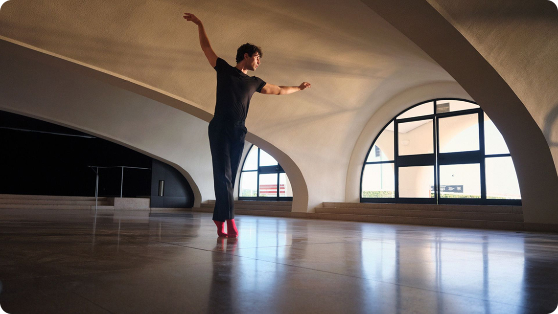 Male dancer in black outfit and red socks balancing on one foot in a spacious room with arched windows.