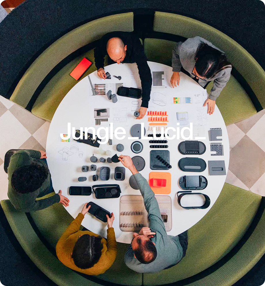 Overhead view of five people seated around a round table covered with various small black and gray product prototypes and design sketches.
