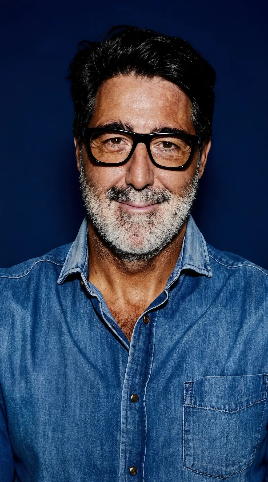 Middle-aged man with black and gray hair and beard wearing black glasses and a blue denim shirt smiling against a dark blue background.
