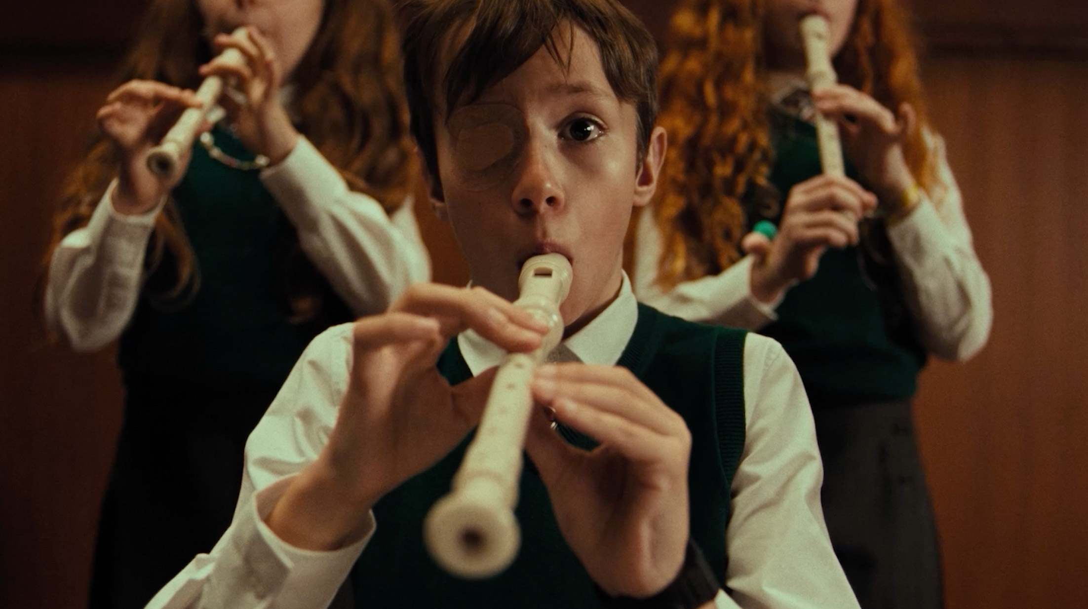 Niño con parche en el ojo tocando una flauta dulce, acompañado por otros dos niños al fondo que también tocan flautas.