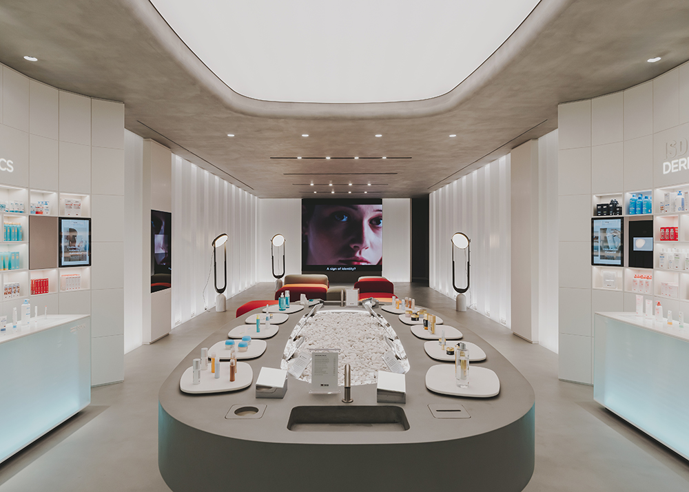 Interior moderno de una tienda de cuidado de la piel con mostradores iluminados, exhibición central de productos y una pantalla grande con el rostro de una persona.