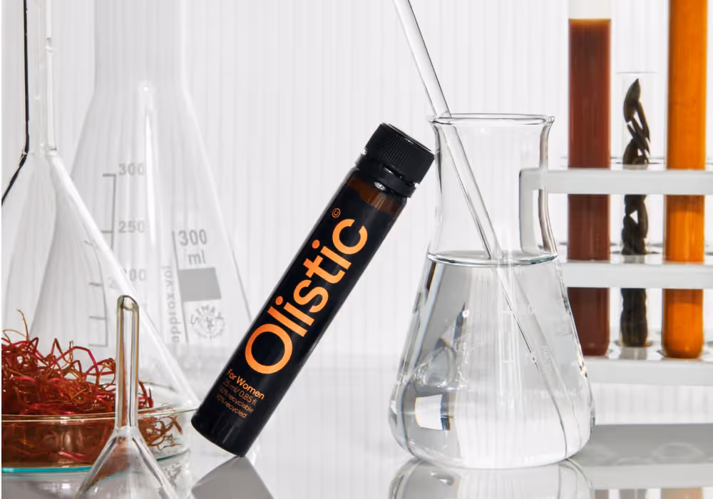 Frasco oscuro con etiqueta Olistic junto a matraces, probetas con líquidos y hierbas en un entorno de laboratorio.
