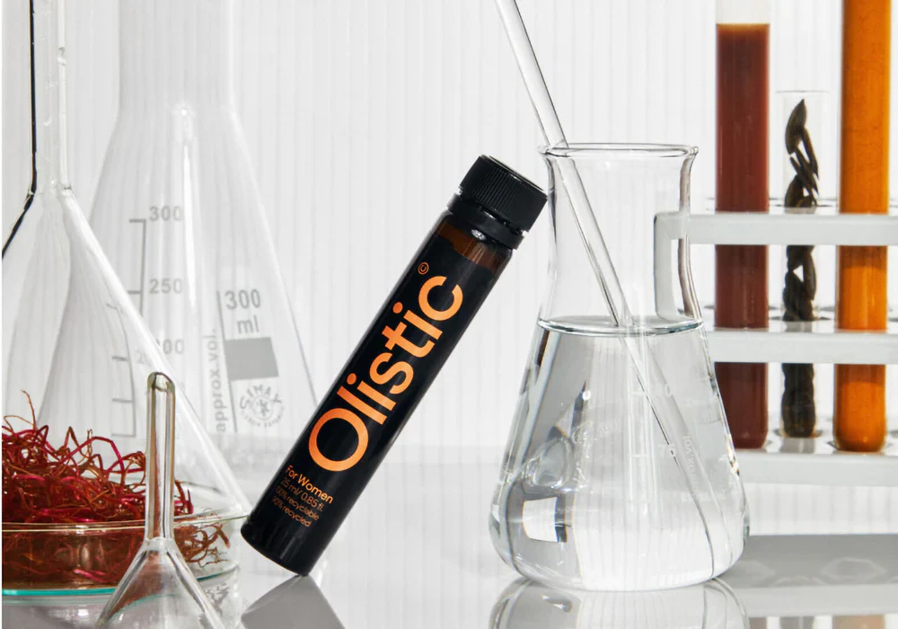 Frasco oscuro con etiqueta Olistic junto a matraces, probetas con líquidos y hierbas en un entorno de laboratorio.
