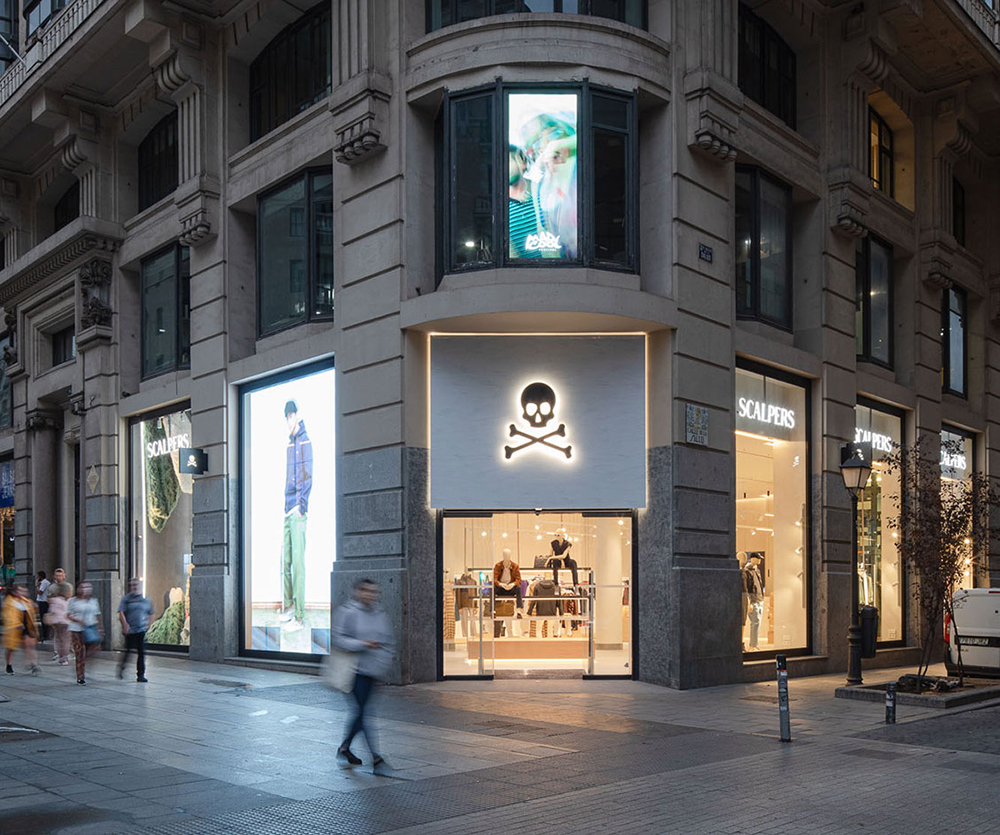 Vista exterior de una tienda Scalpers en un edificio de esquina con logo de calavera iluminado y vitrinas con maniquíes.
