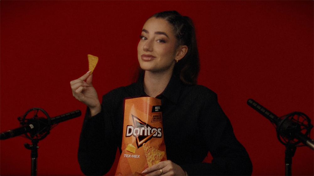 Mujer sosteniendo un triángulo de Doritos mientras sostiene una bolsa de Doritos sabor Tex-Mex frente a un fondo rojo.