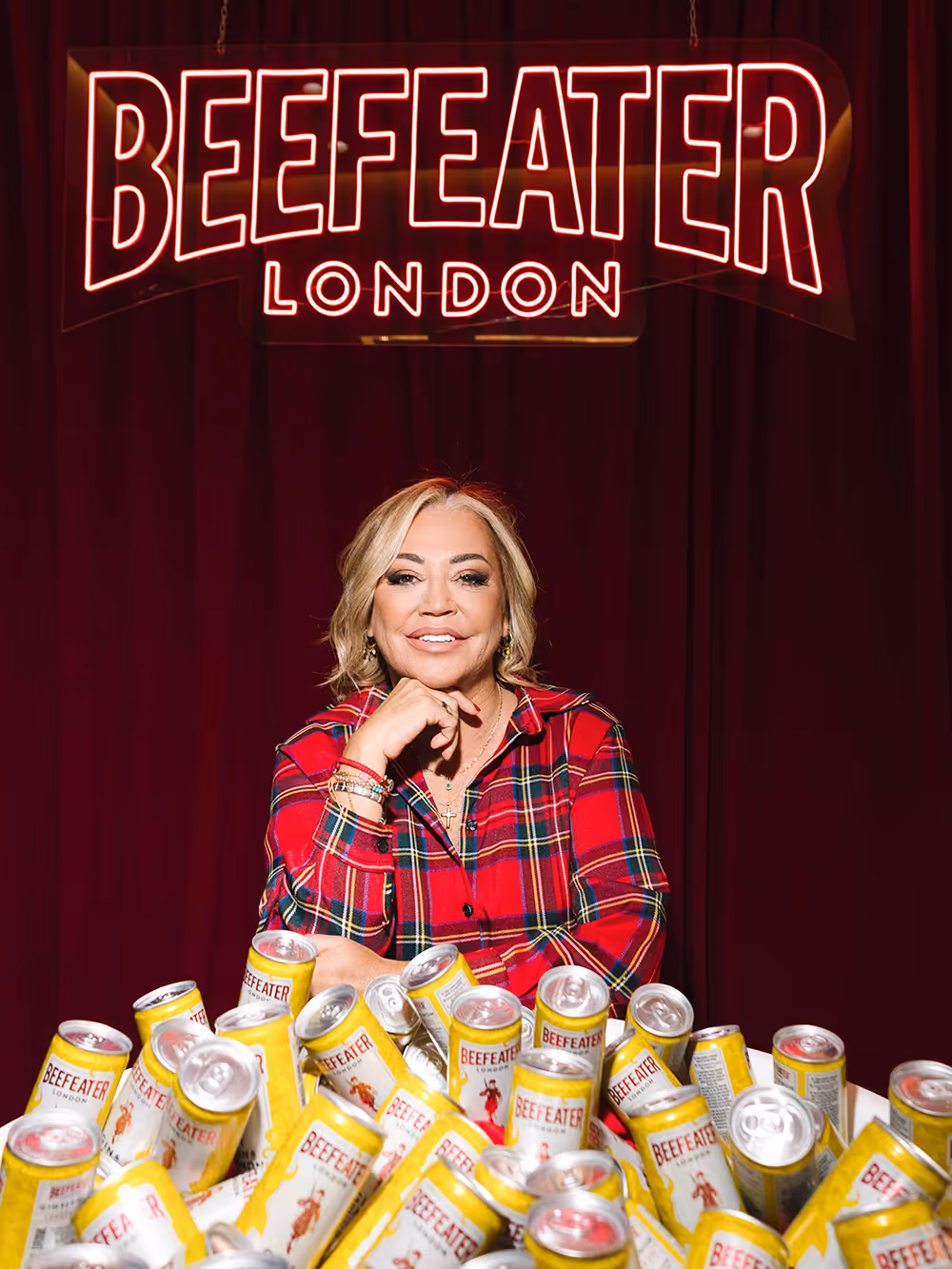 Mujer sonriente con camisa a cuadros rojos frente a un montón de latas de Beefeater bajo un letrero de neón rojo que dice Beefeater London.