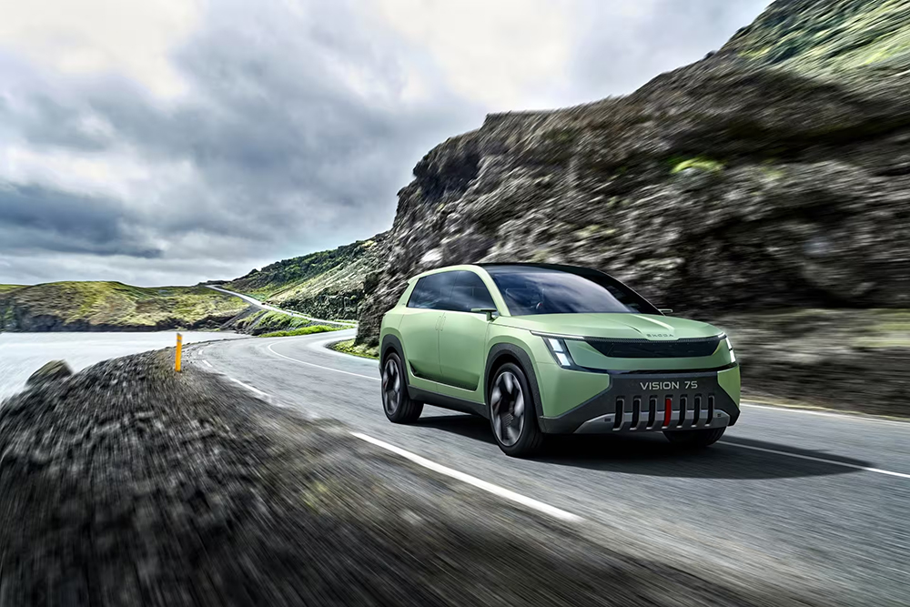 Coche eléctrico verde Škoda Vision 7S circulando por una carretera costera con colinas rocosas y cielo nublado.