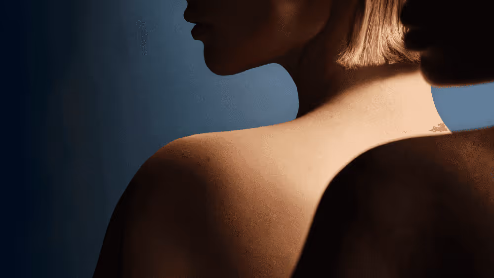 Primer plano del perfil de dos personas con hombros desnudos, una con piel clara y cabello rubio, y otra con piel oscura, sobre un fondo azul oscuro.