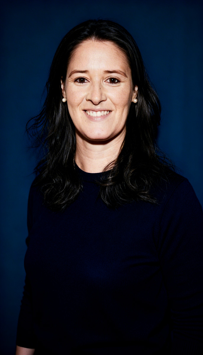 Mujer con cabello oscuro y lacio, vistiendo suéter azul marino y pendientes de perla, sonríe frente a un fondo azul.