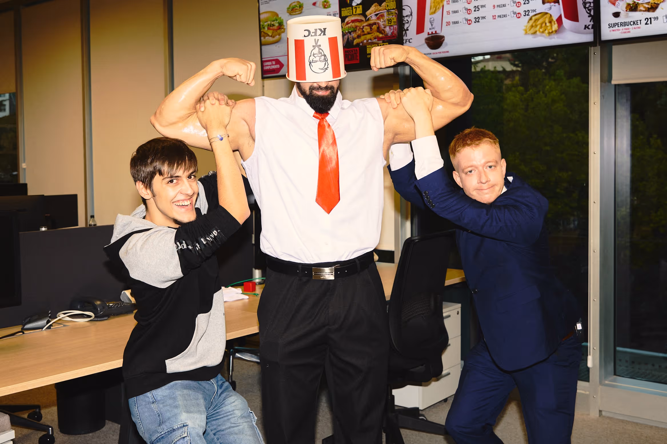 Tres hombres en una oficina imitan a un hombre musculoso con un cubo de KFC en la cabeza mientras muestran fuerza y camaradería.