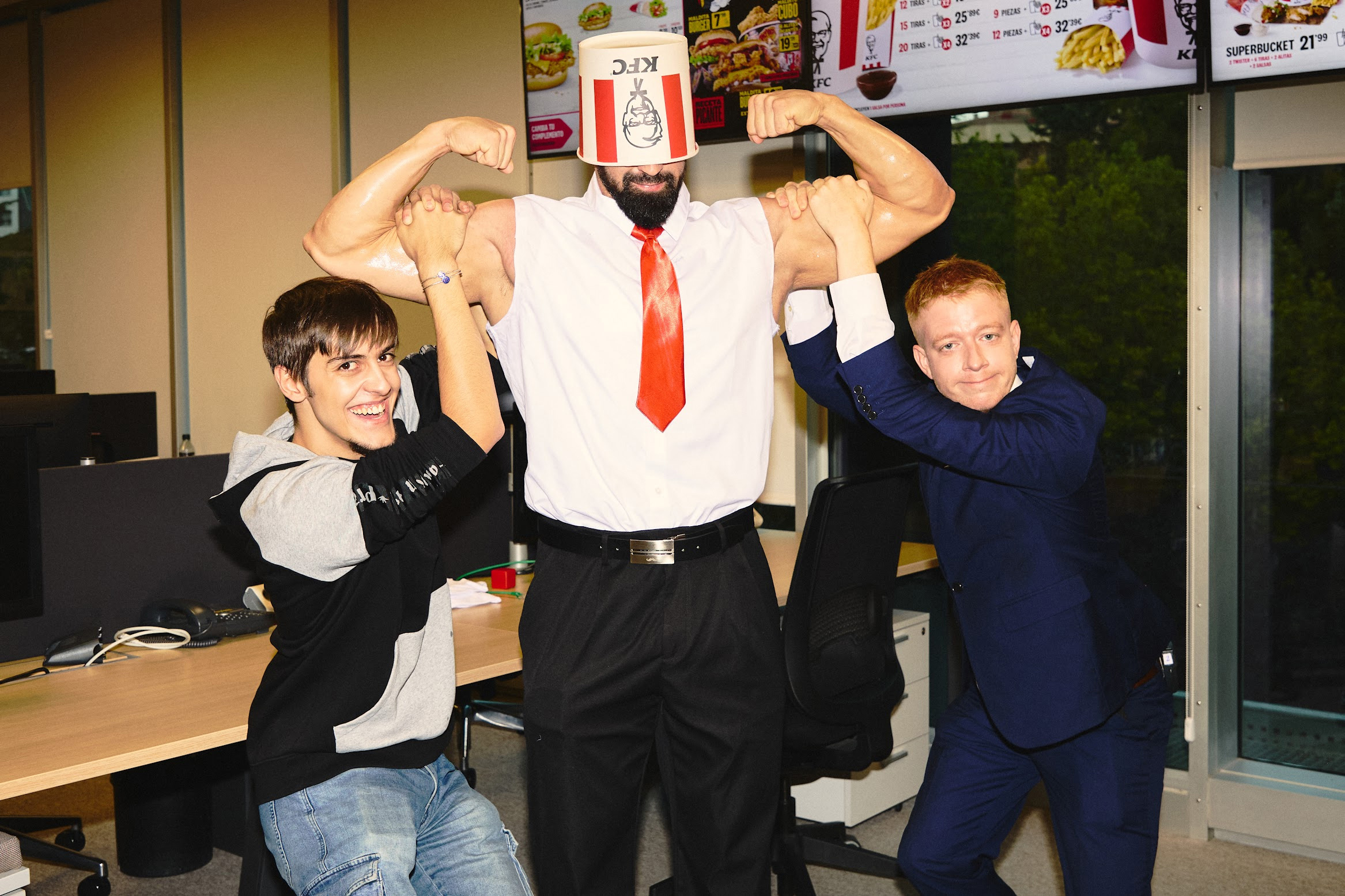 Tres hombres en una oficina imitan a un hombre musculoso con un cubo de KFC en la cabeza mientras muestran fuerza y camaradería.